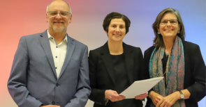 Mann und zwei Frauen stehen lächelnd zusammen, die mittlere hält ein Dokument.