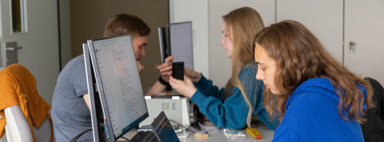 Three students are working at a desk with multiple monitors in a seminar room. A female student in the foreground is typing, while the other two in the background discuss a piece of hardware.