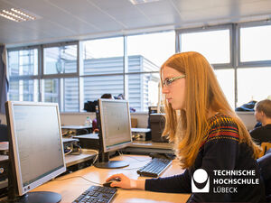 Eine junge Frau sitzt an einem Computer-Arbeitsplatz und arbeitet. Der Arbeitsplatz hat einen Computer-Maus und einen Computer-Keyboard, die auf dem Tisch vor der Frau liegen.