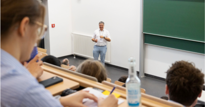 Ein Mann im Hintergrund, steht vor einer Tafel in einem Hörsaal