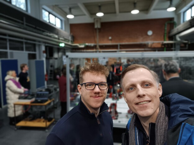 Selfie zweier Besucher im Labor mit Maschinen im Hintergrund.