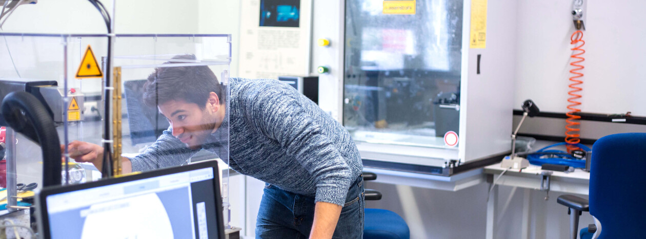 A student leans over to work intently on a piece of technical equipment inside a safety enclosure. A computer screen is visible in the foreground.