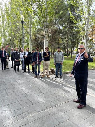 A man in a suit gives a thumbs-up while leading a tour group outdoors.