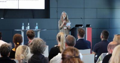 Eine Frau im Hintergrund hält eine Rede, im Vordergrund Publikum