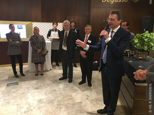 A man in a suit speaks to a small crowd during an indoor reception.
