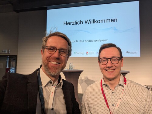 Selfie zweier Konferenzteilnehmer vor Begrüßungsfolie zur 6. KI-Landeskonferenz.