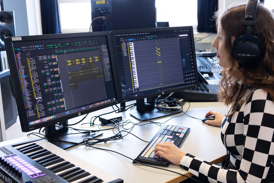 Eine Studierende arbeitet am Computer mit einem Audioschnittprogramm. Die Peripheriegerät bestehen aus Kopfhören, spezieller Tastatur und einem Keyboard. Im Hintergrund ist noch ein Mischpult zu erkennen.