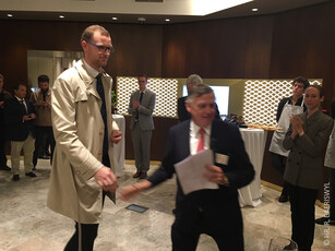A man in a trench coat walks up to shake hands with an award presenter.
