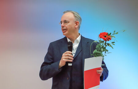 Redner im Anzug mit Mikrofon, Mappe und einer roten Gerbera in der Hand.