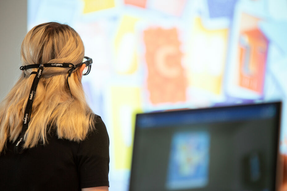 Studierende mit Eye Tracking Brille steht vor einer Leinwand, auf der Bilder gezeigt werden.