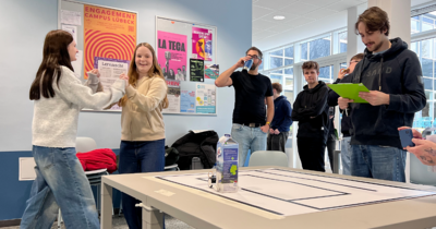 Zwei Schülerinnen albern in einer Pause fröhlich herum, andere schauen zu.