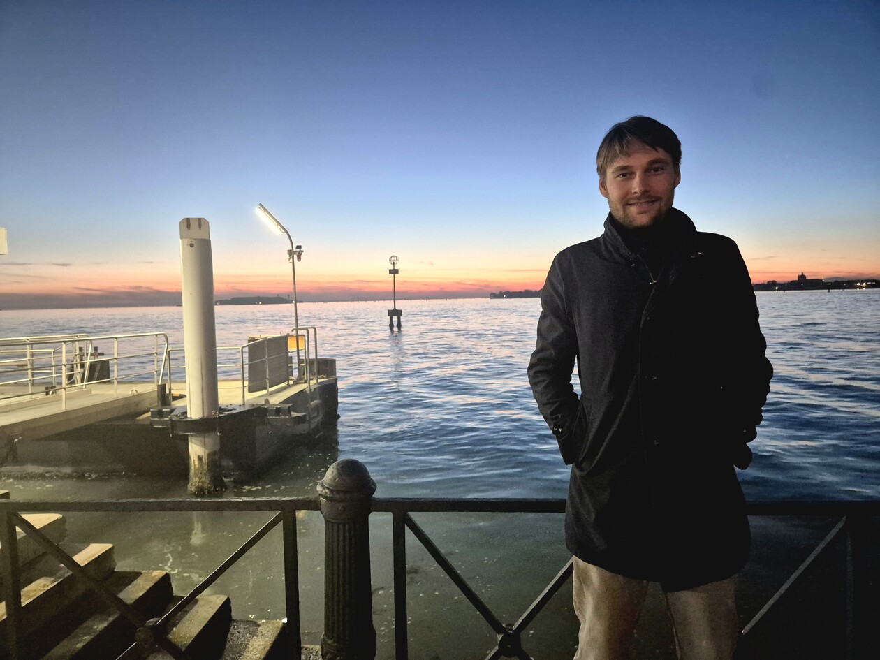 Young man in a dark coat standing by the water during twilight.