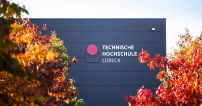 Im Vordergrund Bäume in Herbstfarben, im Hintergrund ein graues Gebäude mit dem Schriftzug TH Lübeck
