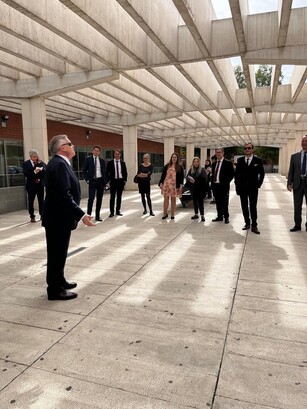 A man in a suit addresses a small group outdoors under a modern pergola.