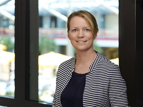 Auf dem Bild sieht man eine Frau mit zurückgebundenen Haaren, blond, mit weiß-blau gestreiften Blazer. 