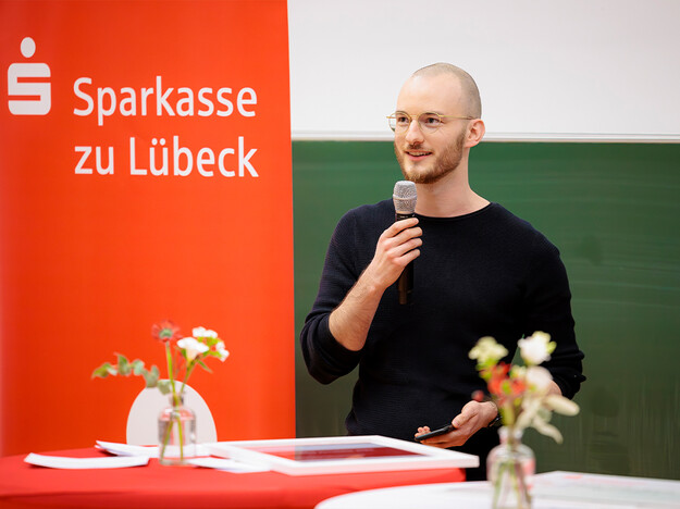 Rede des Gewinnerteams Ein Mann mit einem Mikrofon, neben ihm ein rotes Banner