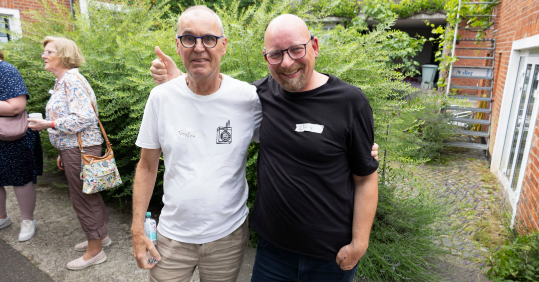 Zwei Männer vor einem grünen Busch, lächeln in die Kamera