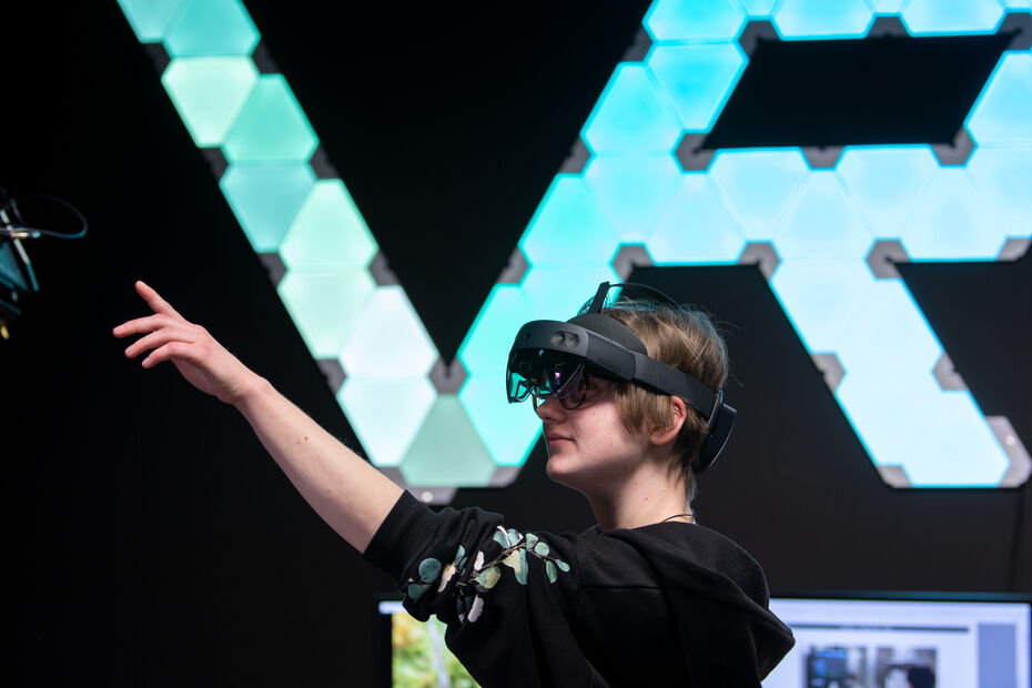 Eine Person mit VR-Headset steht vor einer Wand mit einem leuchtenden VR Schriftzug.