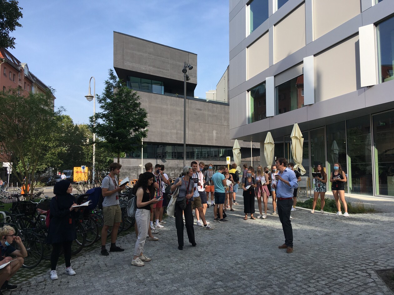 Exkursion vor moderner Architektur: Gruppe hört Erklärungen vor Gebäuden.