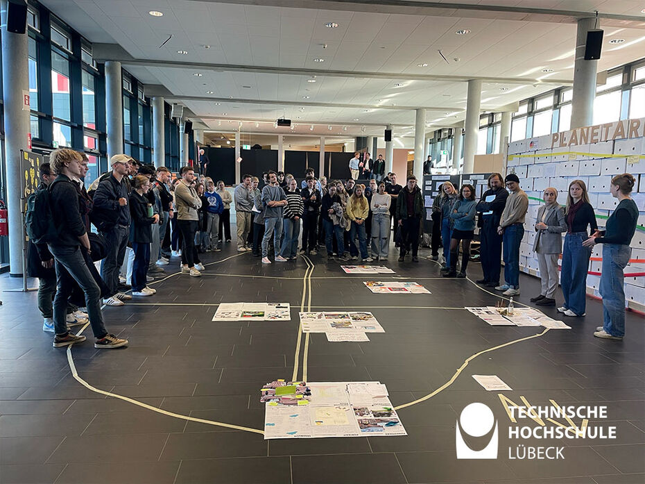 Studierende stehen in UForm im Saal und auf dem Boden befinden sich Plakate die mit Kreppband verbunden sind