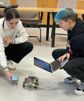 Hörakustik-Studierende testen einen selbstprogrammierten Roboter-Prototypen mit Laptop und Smartphone auf dem Boden des Labors.