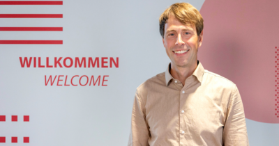 Ein Mann in beigem Hemd steht vor einer Wand auf der Willkommen und Welcome steht. Er lächelt in die Kamera