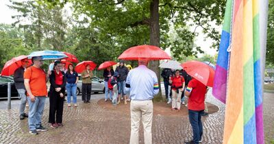 Mehrere Menschen stehen im Halbkreis mit Regenschirmen vor der Flagge