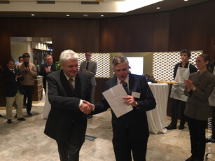 Two men in suits shake hands at a reception as a crowd applauds.