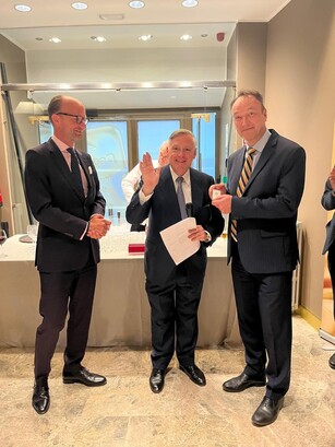 Three men pose for a photo at a reception; the presenter in the middle is waving.