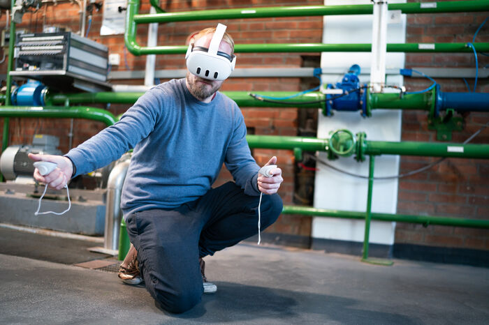 Mann benutzt kniend VR / AR - Brille vor grünen Rohren in einer Werkstatt