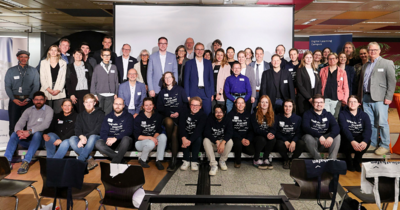 Großes Gruppenfoto auf einer Bühne, viele tragen dunkelblaue Hoodies.