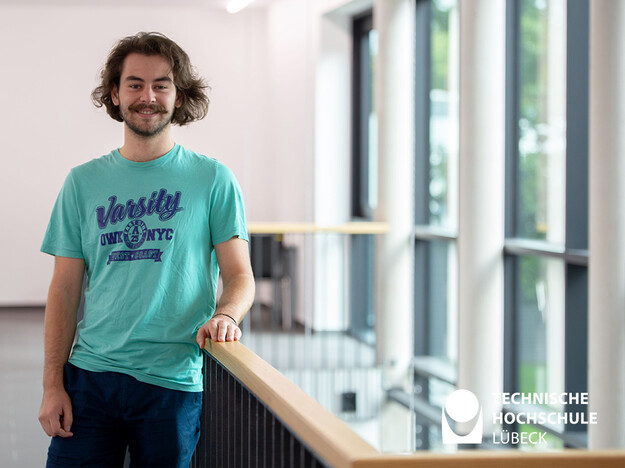 Studieren an der TH Lübeck mit dem Erasmus Programm Ein Mann in türkisem T-Shirt mit mittellangen braunen Haaren und französischen Bart lehnt an einem Geländer