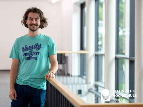 Ein Mann in türkisem T-Shirt mit mittellangen braunen Haaren und französischen Bart lehnt an einem Geländer