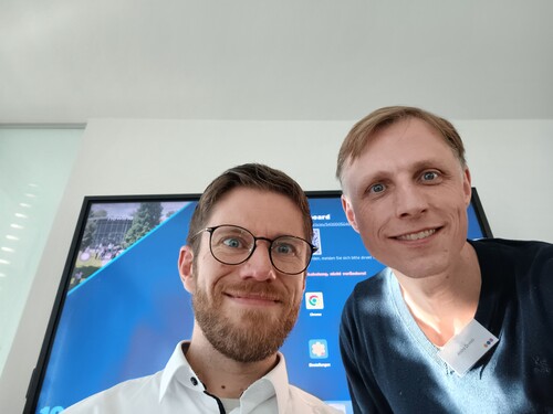 Selfie von zwei Teilnehmenden vor Präsentationsbildschirm.