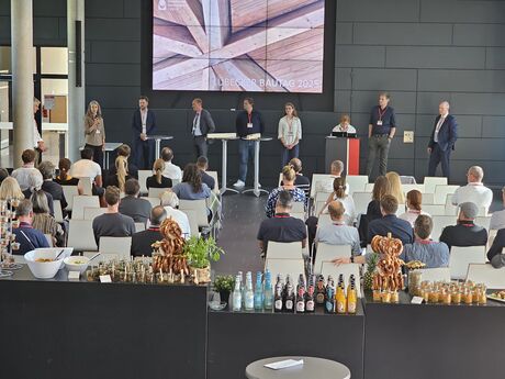 Mehrere Personen stehen und sitzen auf einer Bühne zur Podiumsdiskussion; davor sitzt das Publikum, im Vordergrund ein Buffet mit Getränken und Snacks.