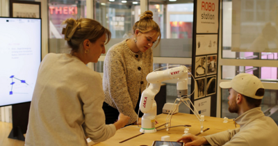 3 junge Menschen stehen und sitzen um eine Station mit einem Roboter herum