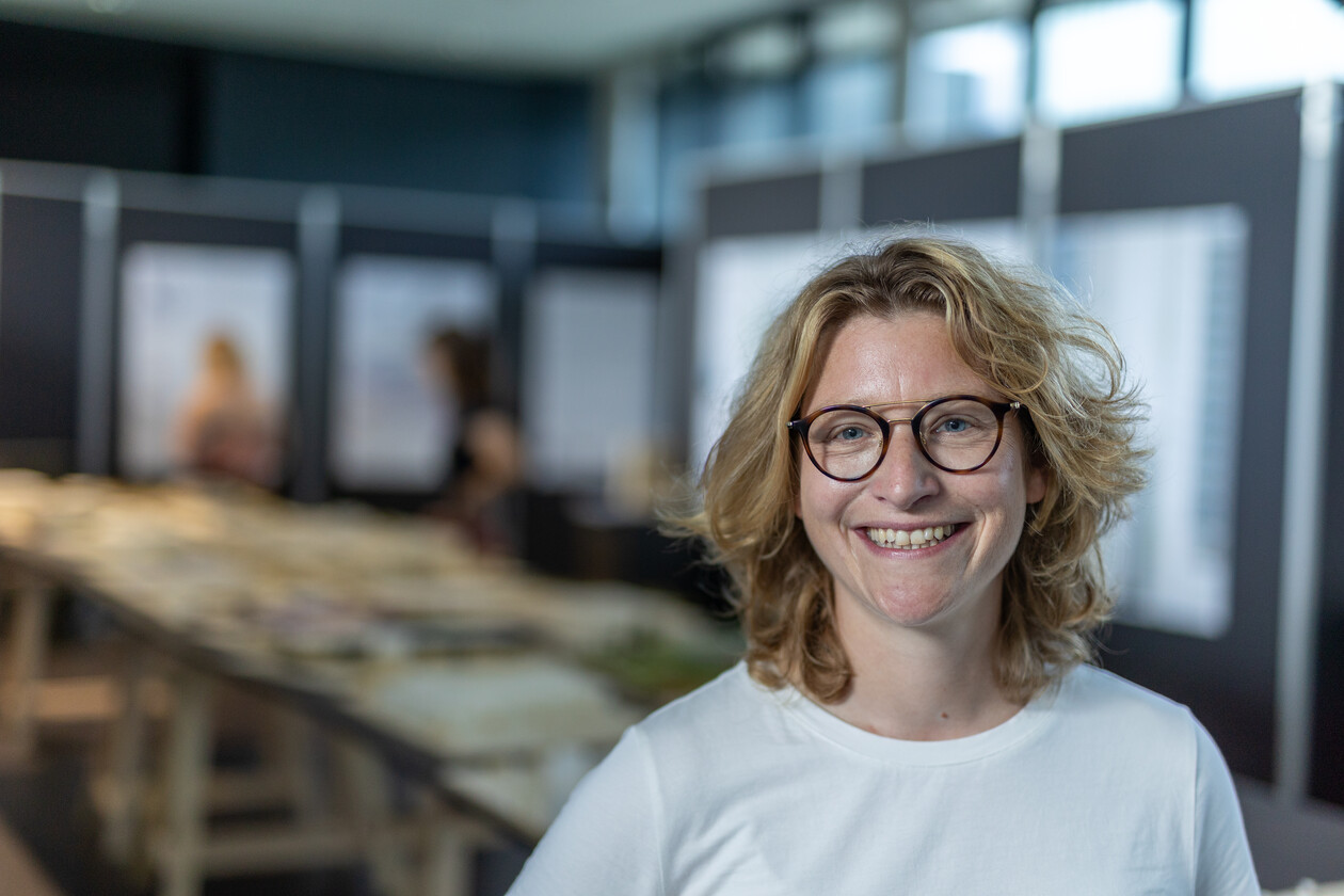 Lächelnde Frau mit Brille und weißem Shirt vor Präsentationstafeln.