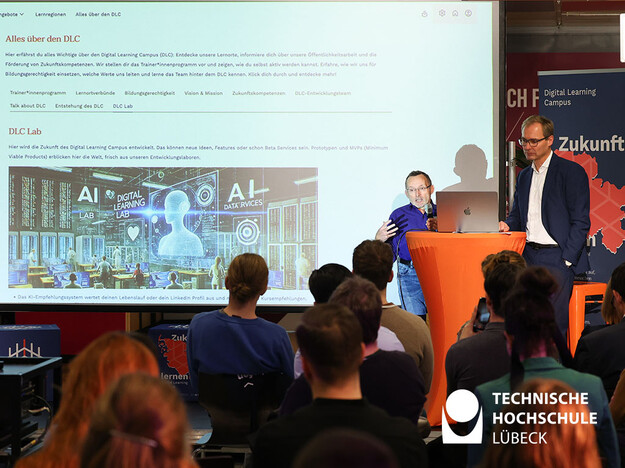 Professor erklärt die Plattform Digital Learning Campus Zwei Männer stehen am Rednerpult und das Publikum blickt zu ihnen
