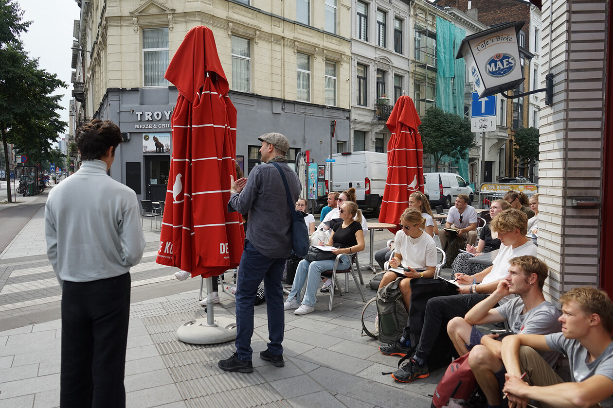Studierende sitzen vor einem Café, zeichnen und hören einem Dozenten zu.