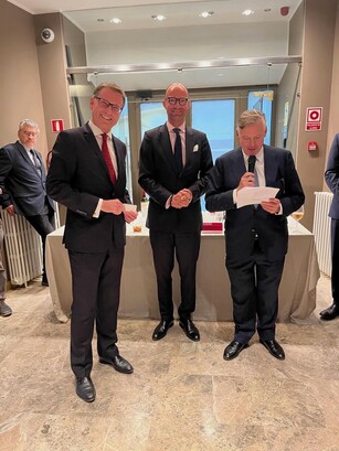 Three men in suits pose at a reception; one is smiling broadly.