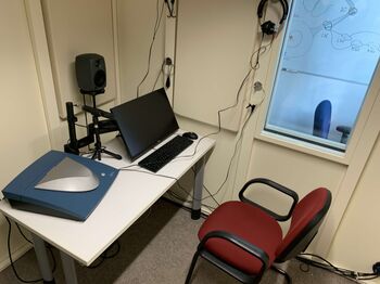 Audiometrie-Arbeitsplatz im Labor mit Computer, 3D-Ohrscanner, Kopfhörern und Sichtfenster zur schallisolierten Messkabine.
