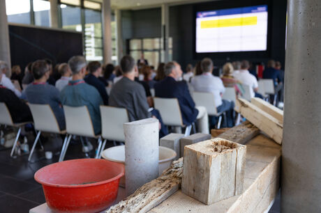 Im Vordergrund liegen Baumaterialien wie Holz und Beton, im Hintergrund sitzt das Publikum und verfolgt eine Präsentation.