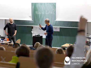 Zwei Professoren der TH Lübeck stehen vor einer Tafel in einem Hörsaal und erklären das Licht. Ein Kind aus dem Publikum hat eine Frage und hebt die Hand.