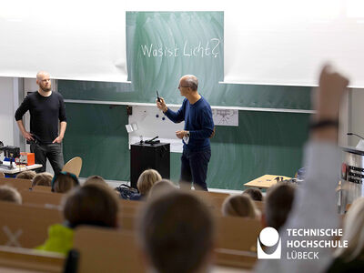 Zwei Professoren der TH Lübeck stehen vor einer Tafel in einem Hörsaal und erklären das Licht. Ein Kind aus dem Publikum hat eine Frage und hebt die Hand.
