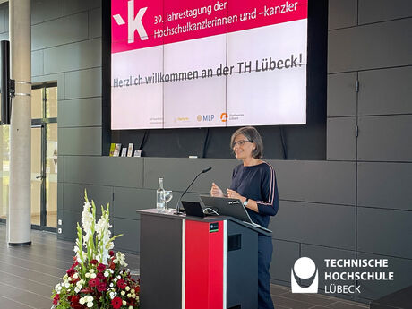 Präsidentin steht am Pult mit Präsentation an Leinwand