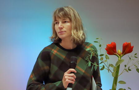 Frau im karierten Pullover spricht ins Mikrofon neben einer Amaryllis.