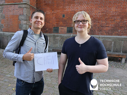 Auf der Suche nach Kommilitonen Zwei Männer stehen nebeneinander mit dem Daumen nach oben und einem Schild in der Hand das sagt "Chemie".