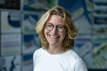 Lächelnde blonde Frau mit Brille vor einer Wand mit vielen Bildern.