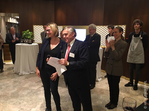 A man with a microphone stands with a female award winner at a reception.