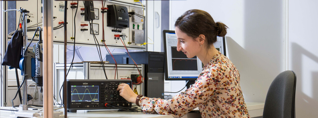 A female student sits at a desk in an electrical engineering lab, operating an oscilloscope that displays a sine wave. In the background is a switchboard with measuring instruments.